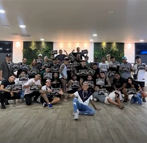 Varney, sentado no chão à frente, com a turma do segundo workshop numa sala clara com paredes de plantas, todos segurando certificados pretos